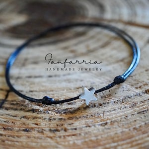 May include: A handmade black cord bracelet with a silver star charm. The bracelet is knotted on each end and features the text "Fanfarria Handmade Jewelry". The bracelet is displayed on a wooden surface.