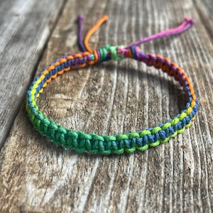 May include: A handmade macrame bracelet in a vibrant array of colors. The bracelet features a woven design with green, blue, yellow, orange, and purple threads. The adjustable closure uses the same colorful threads. The bracelet is displayed on a rustic wooden surface.