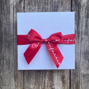May include: A white square gift box tied with a red satin ribbon and bow. The ribbon has the text "For Infarria" in white script. The box is on a weathered wooden surface.