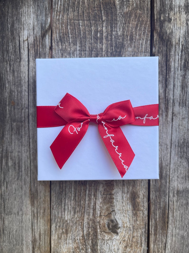 May include: A white square gift box tied with a red satin ribbon and bow. The ribbon has the text "For Infarria" in white script. The box is on a weathered wooden surface.