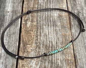 May include: A handmade necklace featuring small, turquoise-colored beads strung on a dark brown cord. The cord is knotted at intervals and has silver-colored accents. The necklace is displayed on a weathered wooden surface.