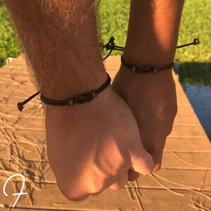 May include: Two dark brown leather bracelets with a knot design and silver accents. The bracelets are worn on wrists. The background shows a wooden surface and green foliage.