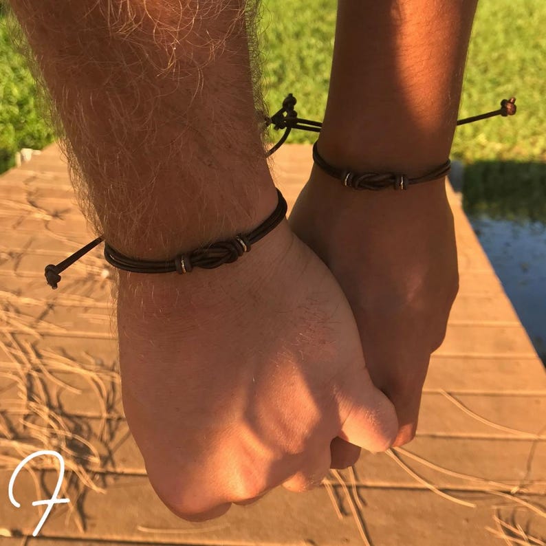 May include: Two dark brown leather bracelets with a knot design and silver accents. The bracelets are worn on wrists. The background shows a wooden surface and green foliage.