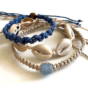 May include: Close-up of several handmade macrame bracelets. One is navy blue with a braided design and a seashell. Another is tan with seashells, and a third is tan with a light blue square bead. The bracelets are on a white surface.