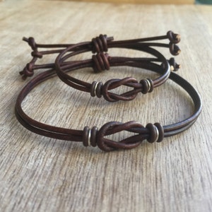May include: Three brown leather bracelets with knot details. The bracelets have a simple design with a knot in the center and silver-colored metal accents. The leather appears to be a rich, dark brown color.
