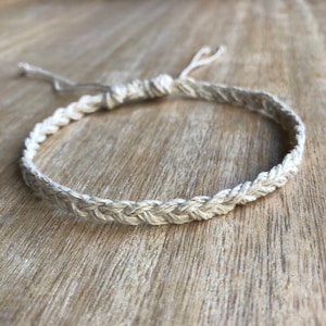 May include: A braided bracelet made of natural-colored twine. The bracelet is adjustable with a sliding knot closure. The woven design is visible, and the bracelet is shown on a wooden surface.