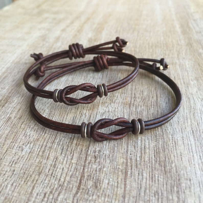 May include: Two brown leather bracelets with a knot design. Each bracelet features a knot in the center and silver-colored metal accents. The bracelets are displayed on a wooden surface.