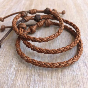May include: Three braided leather bracelets in varying shades of brown. The bracelets have adjustable closures with knotted details. The leather appears to be well-worn, giving them a rustic and natural look. The bracelets are displayed on a light wooden surface.
