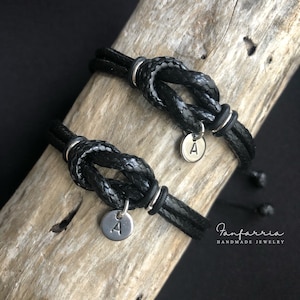 May include: Two black leather bracelets with silver metal knots and a silver charm with the letter A on each. The bracelets are on a wooden surface. The text "Fanfarria Handmade Jewelry" is visible in the bottom right corner of the image.