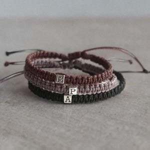 May include: Three macrame bracelets in a stack. The top bracelet is brown, the middle is mauve, and the bottom is black. Each bracelet has a small silver square bead with a letter on it. The letters are B, P, and A.