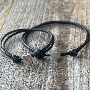May include: Two black cord bracelets with silver heart charms. The bracelets are tied in a knot and the charms are centered on the knot.