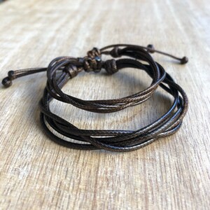 May include: Two brown cord bracelets tied together with a knot. The bracelets are made of thin, braided cord.