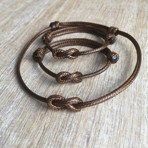 May include: Three brown cord bracelets with infinity knots. The bracelets are layered on top of each other.