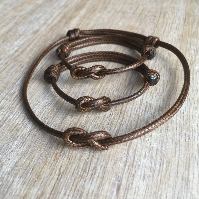 May include: Three brown cord bracelets with infinity knots. The bracelets are layered on top of each other.