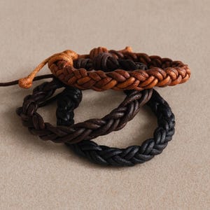 May include: Three braided leather bracelets in shades of brown and black. The top bracelet is light brown, the middle one is dark brown, and the bottom one is black. The bracelets are arranged in a stack.