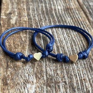 May include: Two blue friendship bracelets with gold heart charms. The bracelets are tied with a knot and are ready to be worn.