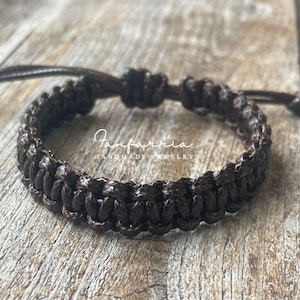 May include: A dark brown braided cord bracelet featuring a woven pattern and adjustable sliding knots. The text Fanfarria HANDMADE JEWELRY is printed in white across the center of the bracelet, which is constructed from a durable, glossy material.