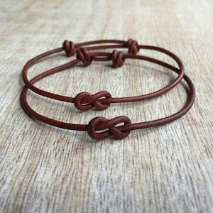 May include: Two brown leather bracelets with a knot design. The bracelets are made of thin, round leather and feature a simple knot closure. The bracelets are displayed on a light brown wooden surface.