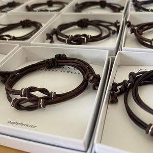 May include: Multiple brown leather bracelets with silver accents are displayed in individual white boxes. Each bracelet features a knot design and is made of multiple strands of leather. The website shopfanfarria.com is visible on the box.