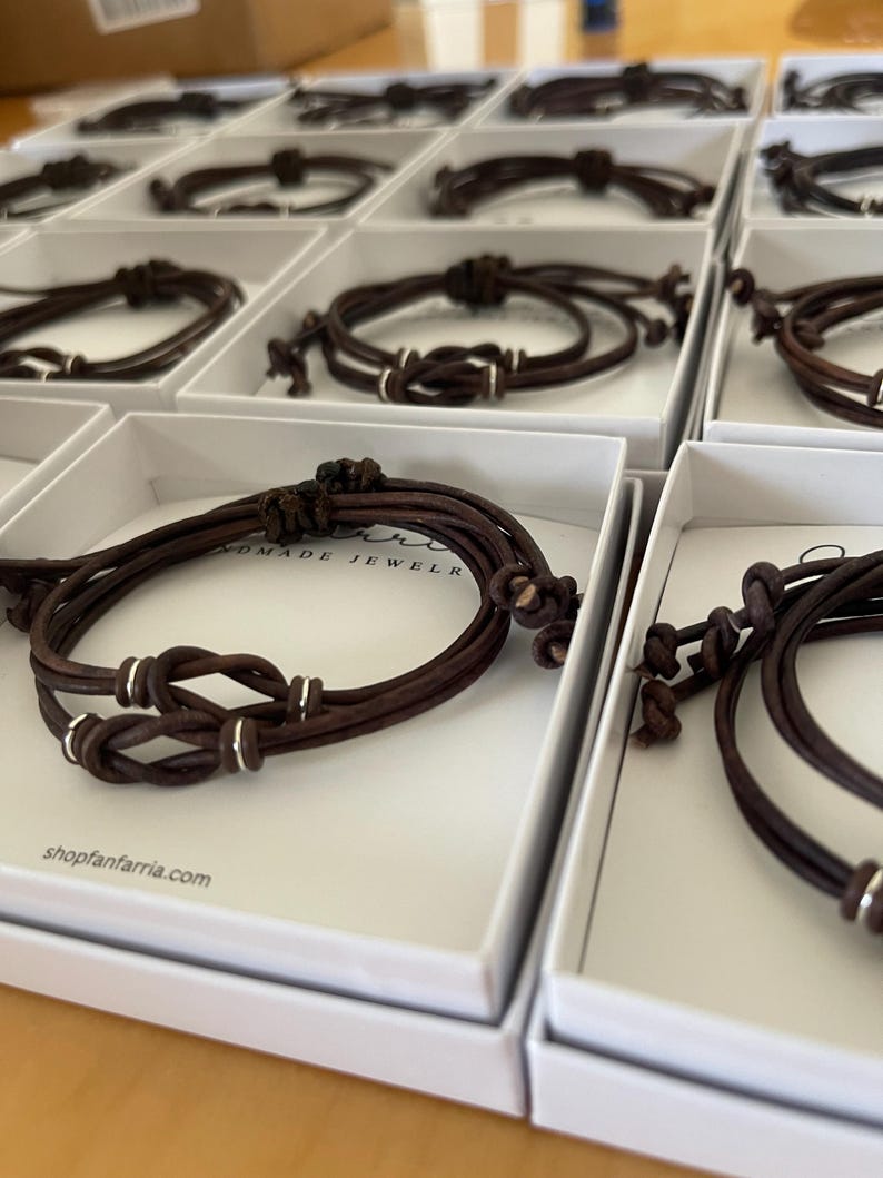 May include: Multiple brown leather bracelets with silver accents are displayed in individual white boxes. Each bracelet features a knot design and is made of multiple strands of leather. The website shopfanfarria.com is visible on the box.