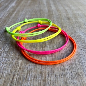 May include: Four neon colored friendship bracelets in yellow, green, pink, and orange. The bracelets are made of thin cord and are tied in a knot.