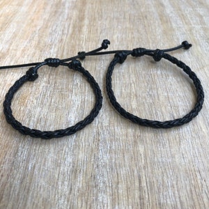 K&ouml;nnte beinhalten: Zwei schwarze geflochtene Lederarmb&auml;nder mit verstellbaren Knoten. Die Armb&auml;nder liegen auf einer Holzoberfl&auml;che.