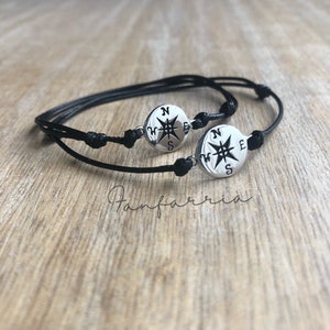 May include: Two black cord bracelets with silver compass charms. Each charm has a compass rose with the letters N, S, E, and W around it.