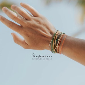 May include: A hand wearing a stack of colorful string bracelets. The bracelets are made of different colors, including green, yellow, orange, and beige. The bracelets are tied with a knot at the wrist. The text "Fanfarria Handmade Jewelry" is visible at the bottom of the image.