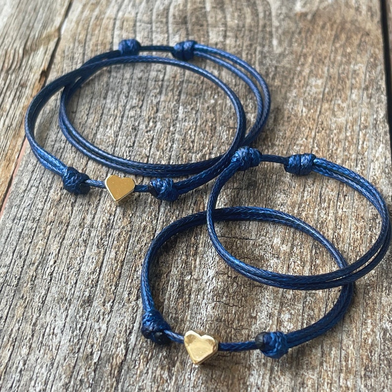May include: Two navy blue friendship bracelets with gold heart charms. The bracelets are tied together in a knot.