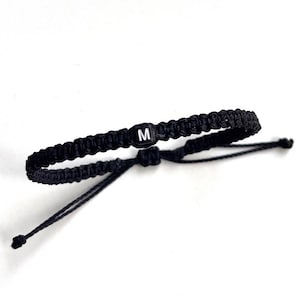 May include: Black braided bracelet with an adjustable sliding knot closure. The bracelet features a small, square bead with the letter "M" in white. The bracelet is shown against a white background.