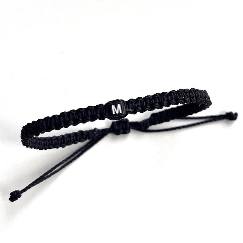 May include: Black braided bracelet with an adjustable sliding knot closure. The bracelet features a small, square bead with the letter "M" in white. The bracelet is shown against a white background.