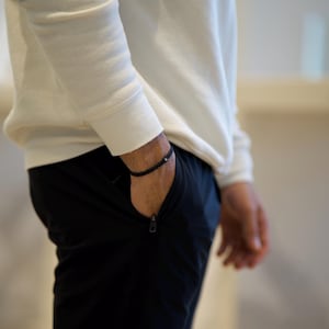 May include: A black beaded bracelet is worn on the wrist. The person is wearing a white long-sleeve shirt and black pants with a zipper pocket. The image is a medium shot.