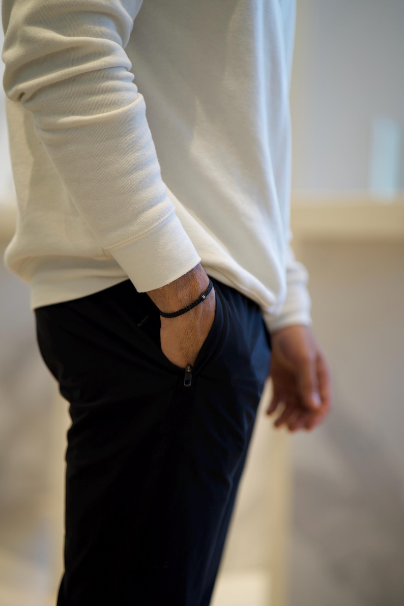 May include: A black beaded bracelet is worn on the wrist. The person is wearing a white long-sleeve shirt and black pants with a zipper pocket. The image is a medium shot.