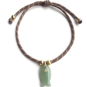 May include: A brown cord bracelet featuring a light green jade-like fish pendant. The cord has white diagonal stripes and is secured with a sliding knot. Two gold-toned metal beads flank the pendant, which is accented with a small clear stone.