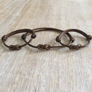 May include: Three brown friendship bracelets with infinity knots. The bracelets are made of a thin, braided cord.