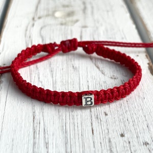 May include: A red macrame bracelet with a silver-colored square bead featuring the letter "B". The bracelet is adjustable with sliding knots. The bracelet is displayed on a white wooden surface.