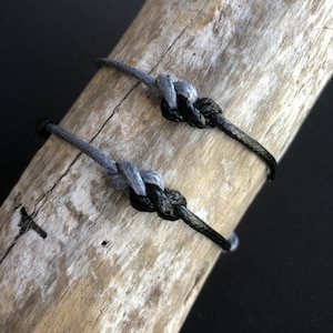 May include: Two bracelets made of knotted cord, one gray and one black, are displayed on a piece of weathered wood.