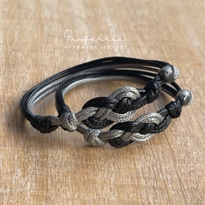 May include: Two friendship bracelets, one black and one gray, are tied together in a knot. The bracelets are made of braided cord and have a simple, minimalist design. The bracelets are on a wooden surface.