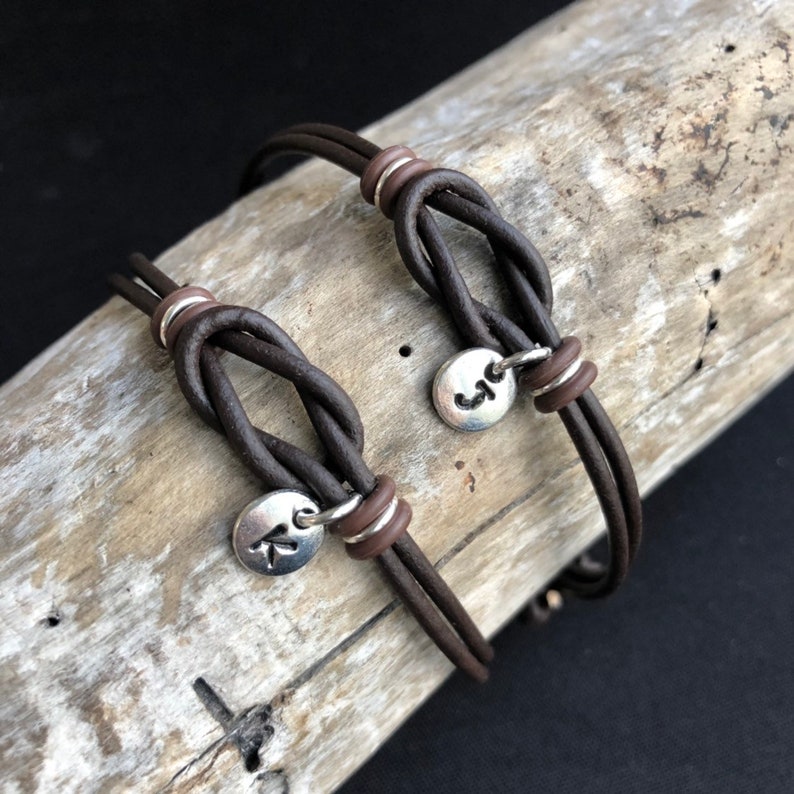 May include: Two brown leather bracelets with a knot design. Each bracelet features a small silver charm with a letter. The bracelets are displayed on a piece of driftwood against a dark background.