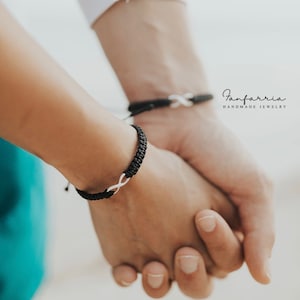 May include: Two people holding hands, each wearing a black string bracelet with a silver infinity symbol charm. The bracelets are from the brand "Fanfarria Handmade Jewelry".