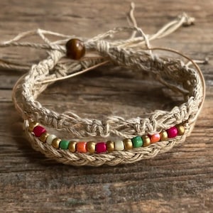 May include: Handmade macrame bracelet with a natural, woven design. The bracelet features a row of colorful beads in red, green, orange, and gold. A wooden bead accents the adjustable closure. The bracelet is displayed on a wooden surface.