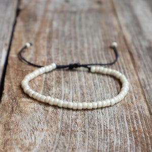 May include: A bracelet featuring a single strand of small, smooth, off-white beads strung on a thin black cord. The design includes an adjustable sliding knot closure with two loose cord ends extending from the back of the circular bead arrangement.