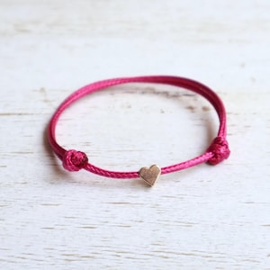May include: A fuchsia pink cord bracelet featuring a small, polished gold-tone metal heart charm centered on the string. The bracelet is constructed from a thin, braided cord with two adjustable knots on either side of the central heart element.