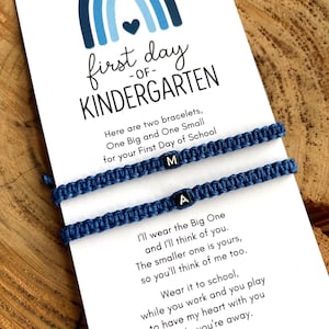May include: A card with the text "first day of KINDERGARTEN" and a blue rainbow graphic. Two blue macrame bracelets with the letters "M" and "A" are displayed. The card includes a poem about the bracelets.