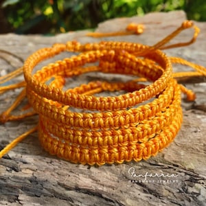 May include: Close-up of several handmade, orange macrame bracelets. The bracelets are stacked on top of each other, showcasing the intricate knotting detail. The bracelets are made of a thick, durable cord. The background is a natural, textured surface.