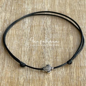 May include: Black cord bracelet featuring a small, detailed silver-colored turtle charm. The bracelet is knotted at each end and has the text "Fanfarrina Handmade Jewelry" in a cursive font.