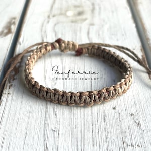 May include: A brown and beige braided bracelet with a red accent. The bracelet is made of natural fibers and has a simple, minimalist design. The text "Fanfarria Handmade Jewelry" is visible on the bracelet.