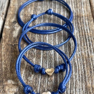 May include: Three blue friendship bracelets with gold heart charms. The bracelets are tied in a knot and are layered on top of each other.