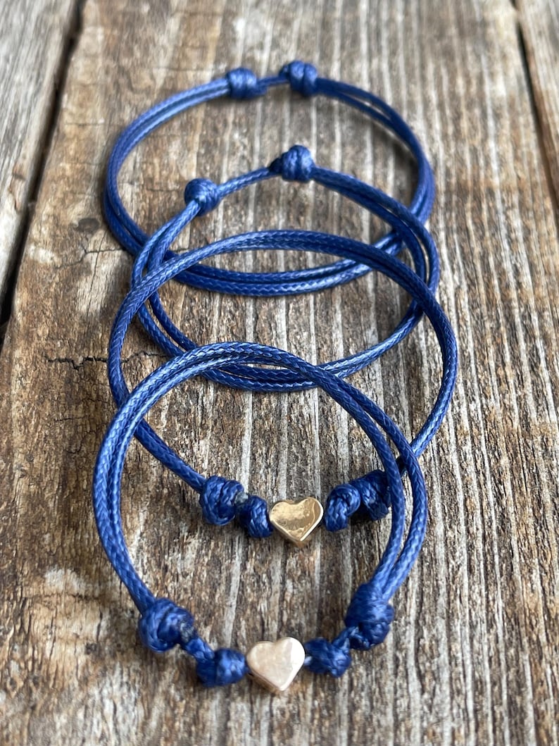 May include: Three blue friendship bracelets with gold heart charms. The bracelets are tied in a knot and are layered on top of each other.