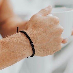 May include: Black braided bracelet with a small white charm featuring a black letter "A". The bracelet is worn on a wrist. The background is blurred, suggesting a casual setting.